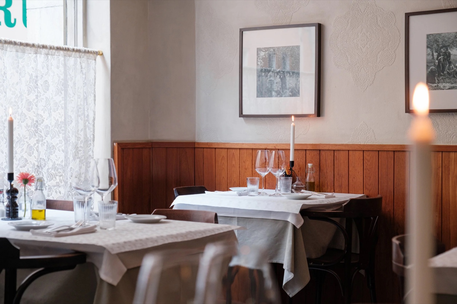 Två dukade bord vid fönstret med tända ljus, vinglas och spetsgardin